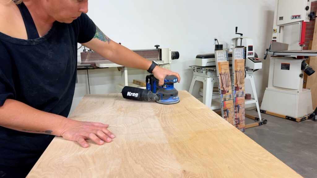 Sanding the plywood workbench top with a random orbital sander for a smooth finish.