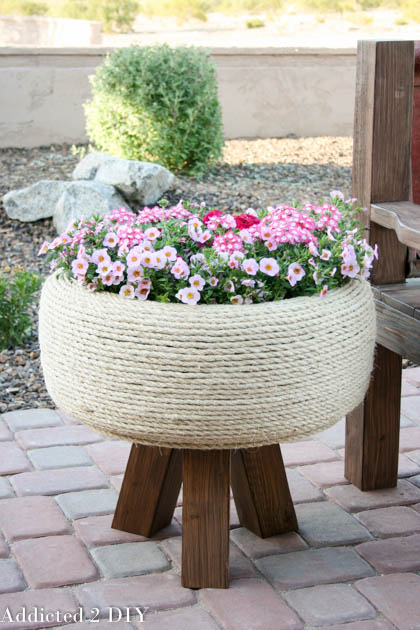 DIY tire planter wrapped in sisal rope and filled with pink flowers on wooden legs