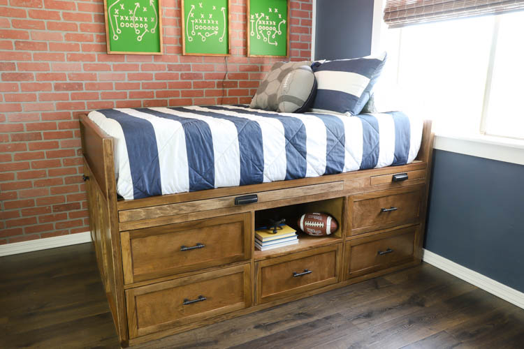 Front view of a finished DIY full-size captain's storage bed with drawers, open shelf, and pull-out nightstand in a boy's sports bedroom