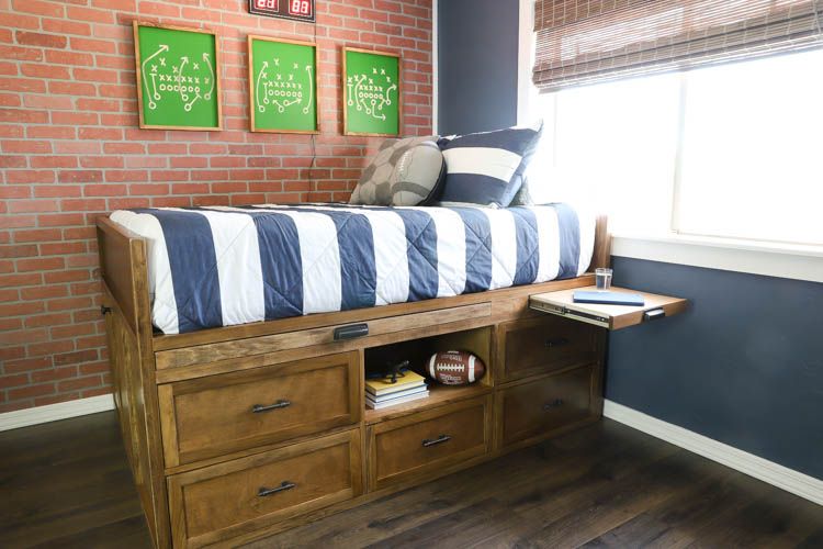 Side view of a finished DIY full-size captain's bed with walnut-stained storage drawers and open cubby in a boy's bedroom with brick accent wall