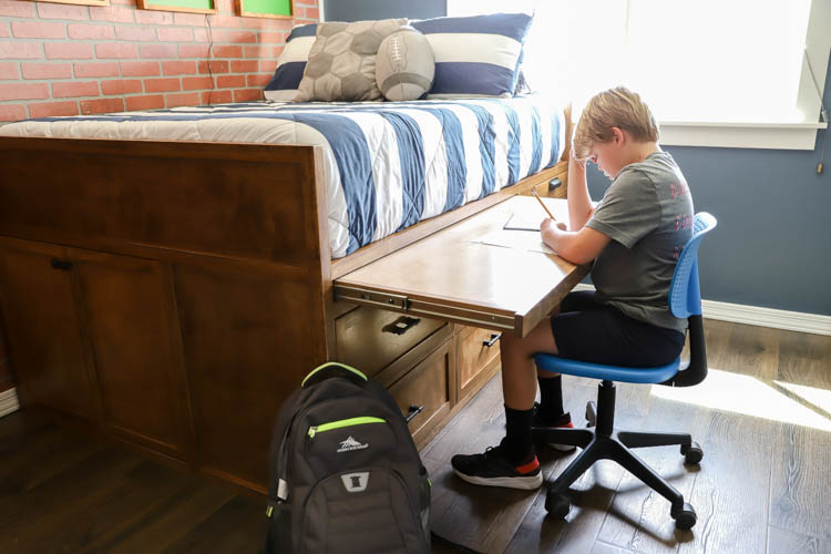 Boy doing homework at the built-in pull-out desk of a DIY full-size captain's storage bed in a sports-themed bedroom