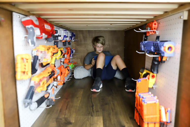 Boy sitting inside the hidden compartment of a DIY captain's bed, surrounded by Nerf guns hanging on pegboard walls