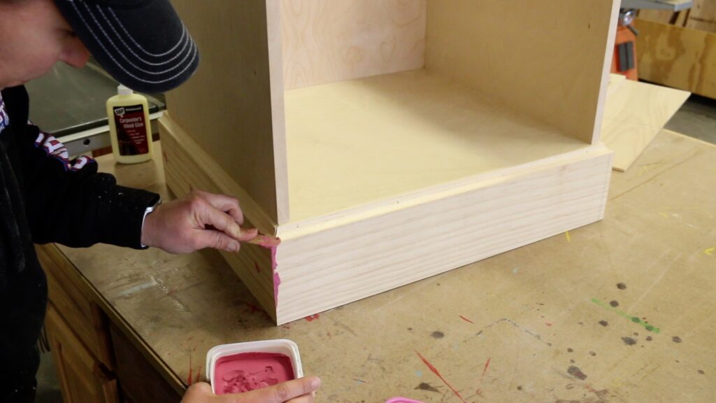 Applying pink DAP Plastic Wood X wood filler to the miter joints on a DIY printer cabinet base
