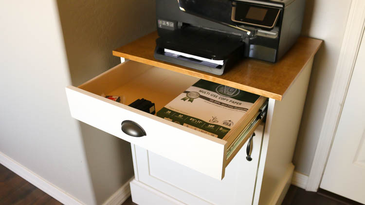 Open drawer of a DIY farmhouse printer cabinet showing paper and ink cartridge storage below a walnut stained cabinet top