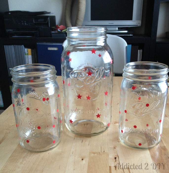Three clear Ball wide-mouth mason jars on a wood workbench with small red vinyl dot and star decals applied randomly across the surface as a paint resist before painting.