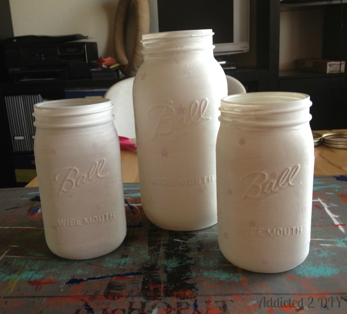 Three Ball mason jars — one half-gallon and two quart-sized — all coated in white Americana Decor Chalky Finish paint as a base layer before adding color, with faint vinyl resist shapes visible.