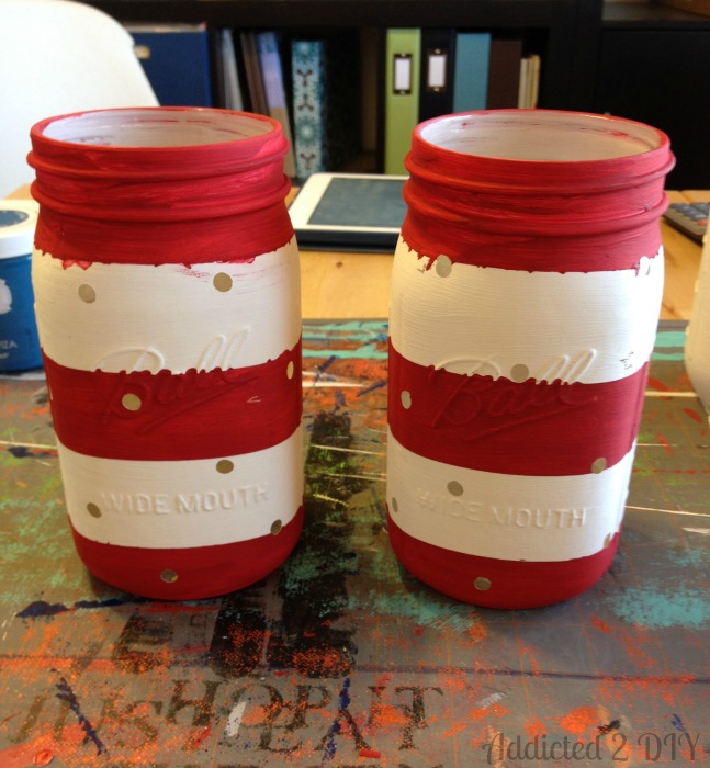 Two quart-sized Ball mason jars painted with red and white horizontal stripes using Frog Tape, with small dot cutouts visible where vinyl resist was removed.