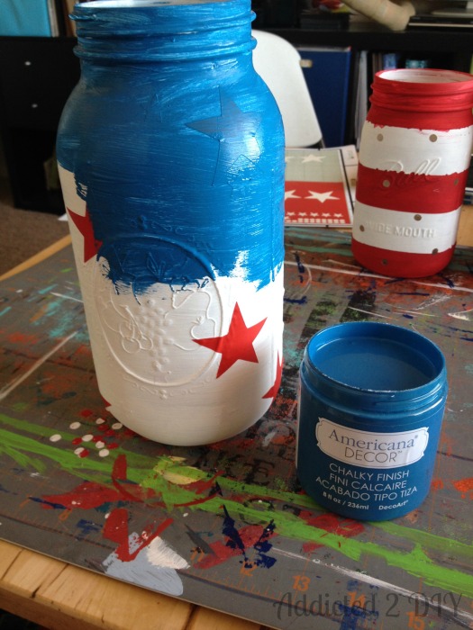 Half-gallon Ball mason jar being painted blue with a red vinyl star resist still in place, next to an open jar of Americana Decor Chalky Finish paint in Legacy.