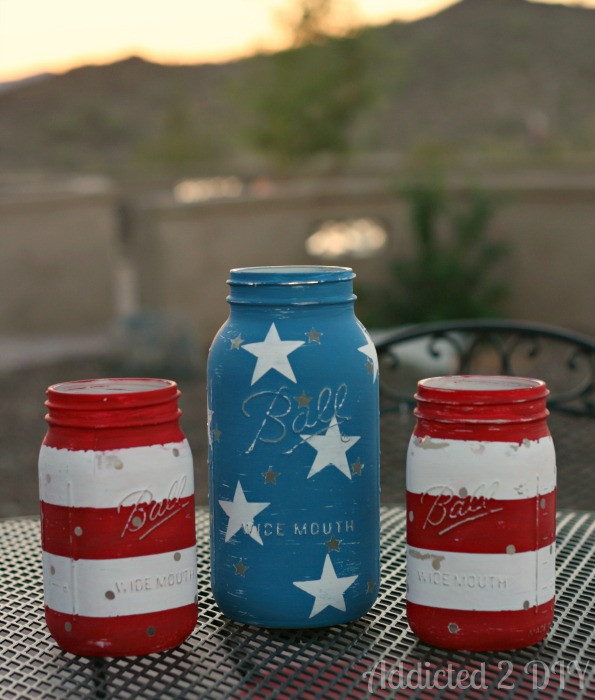 Three completed patriotic mason jar lanterns on an outdoor patio table in daylight — two red and white striped quart jars flanking a distressed blue half-gallon jar painted with white stars.