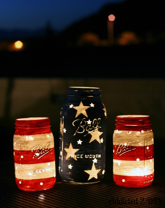 Three chalk painted Ball mason jars glowing with tea lights on a patio table at night, showing red and white striped quart jars and a navy blue half-gallon jar with white stars.