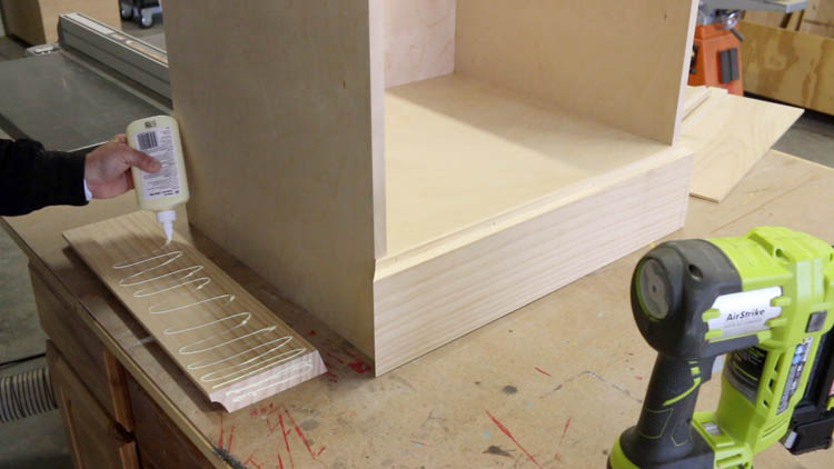 Applying wood glue to a pine moulding board before attaching it to the base of a DIY printer cabinet