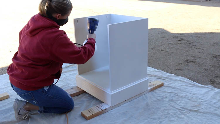 Spray painting a DIY printer cabinet base white using a paint sprayer outdoors on a drop cloth