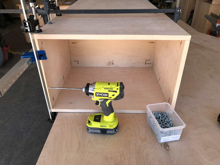 Partially assembled plywood coffee table frame clamped with bar clamps, showing the interior shelf in place, with a Ryobi drill sitting inside