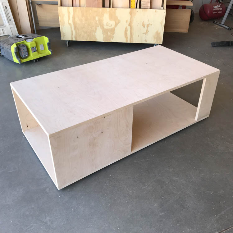 Fully assembled natural plywood coffee table frame with an open lower shelf, sitting on a concrete garage floor before sanding and staining