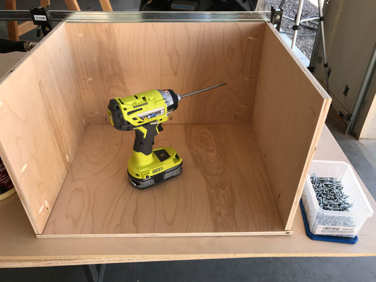 View from inside the coffee table drawer looking down at a Ryobi impact driver being used to secure the bottom panel, with a container of screws beside it