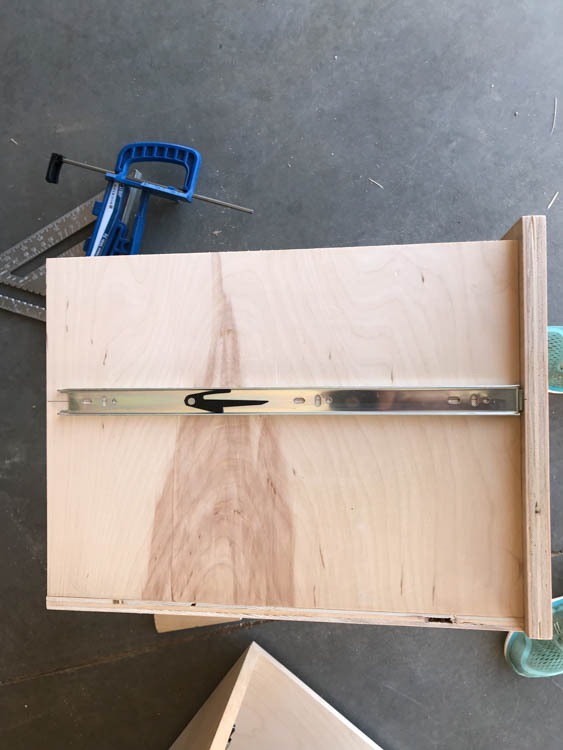 Metal ball-bearing drawer slide laid across the bottom of a plywood drawer box for positioning before installation