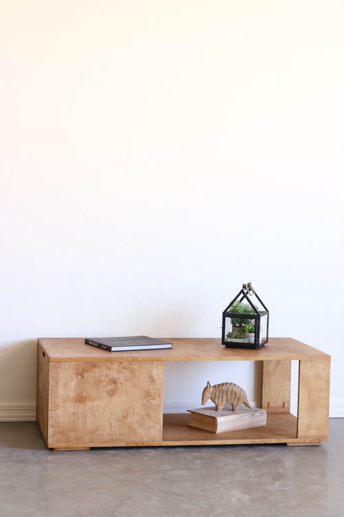 Full-length lifestyle photo of a finished DIY modern coffee table with warm wood stain against a white wall, styled with a coffee table book, glass terrarium, and small armadillo figurine on the lower shelf