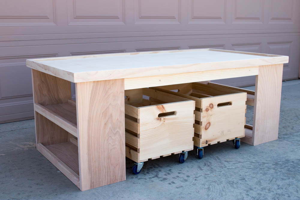 Completed but unpainted DIY kids activity table in natural plywood with two wooden storage crates on casters in a garage