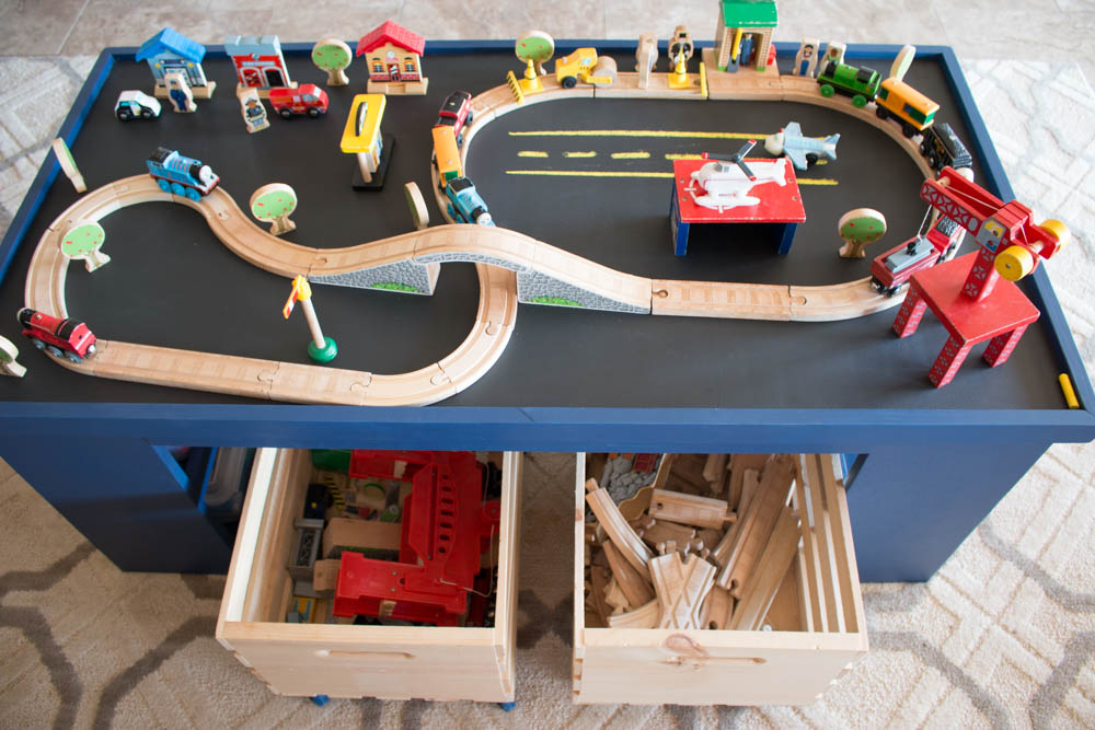 Overhead view of a DIY kids activity table chalkboard side set up with a wooden train track layout and two open storage crates below filled with train tracks