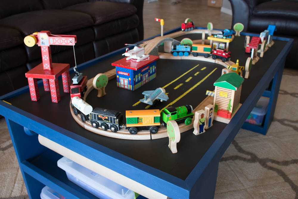 Wooden train set arranged on the chalkboard surface of a navy blue DIY kids activity table with plastic storage bins visible on the side shelves
