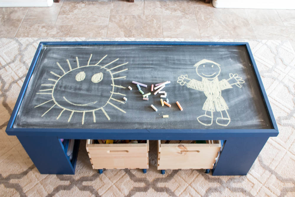 Overhead view of the chalkboard side of a DIY kids activity table with chalk drawings of a sun and a person, surrounded by colorful chalk pieces