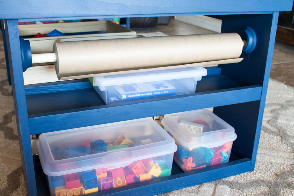 Side view of a navy blue DIY kids activity table showing the built-in drawing paper roll on a dowel, a tray of washable markers, and two plastic bins of Lego bricks on the lower shelf