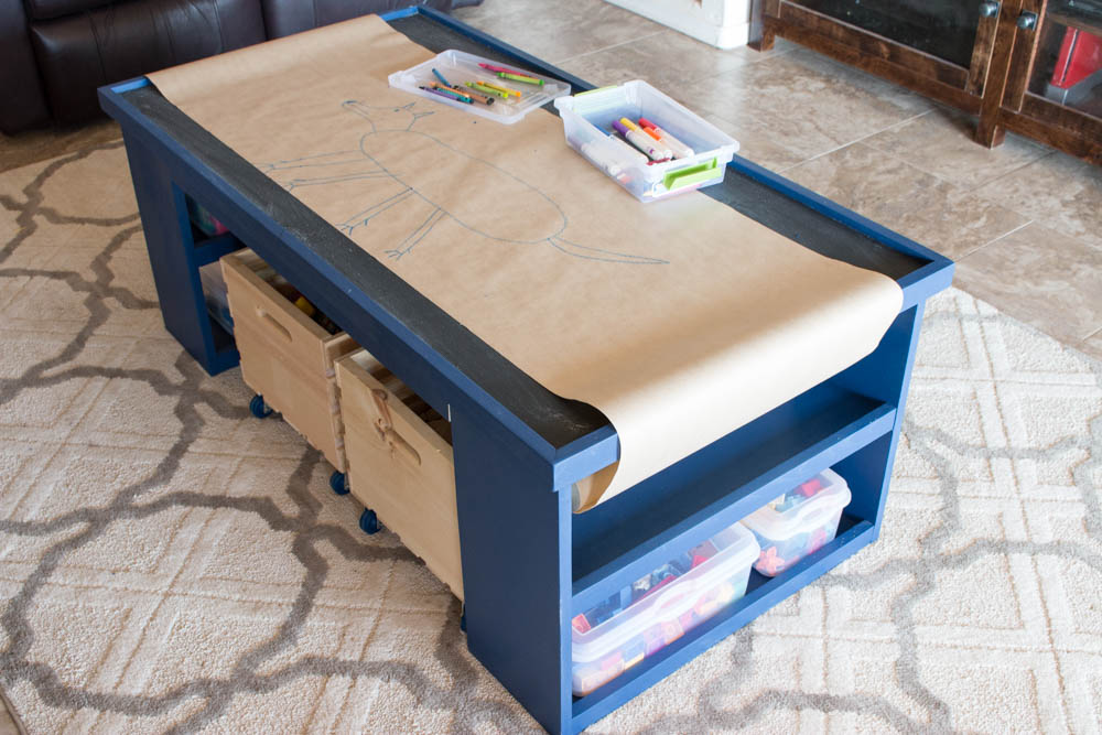 DIY kids activity table with kraft drawing paper rolled out across the chalkboard surface, with crayon and marker bins on top and storage crates and bins below