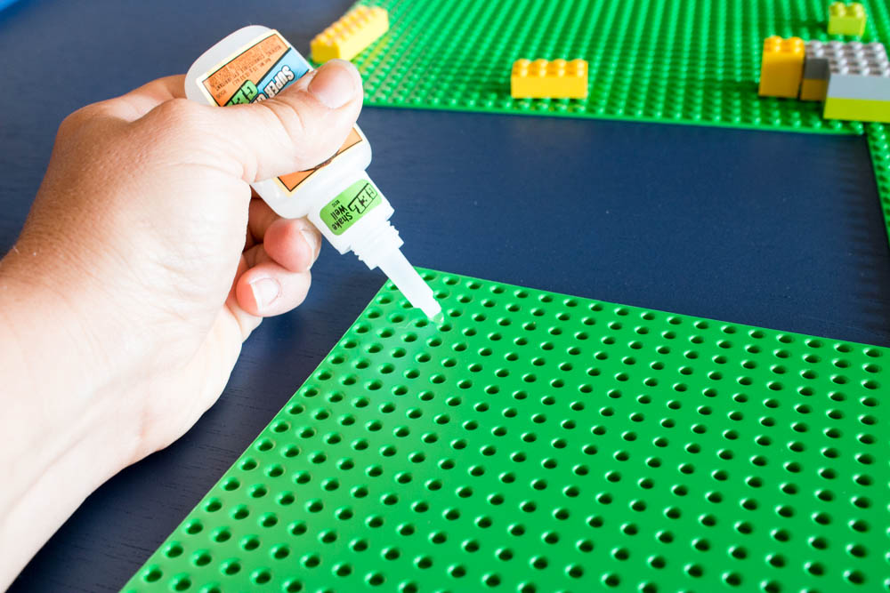 Hand applying super glue to a green Lego base plate on the surface of a DIY kids activity table during assembly