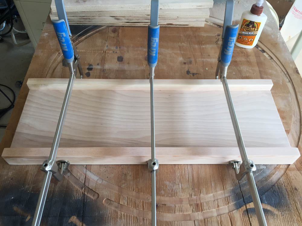 Three Kreg clamps holding a 1x2 board flush to the bottom of a shelf during assembly of a DIY kids activity table with Gorilla Wood Glue visible nearby