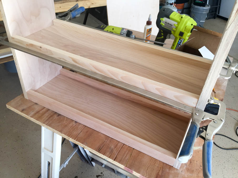 Front view of the DIY kids activity table base mid-assembly showing the bottom and middle shelves attached between the vertical side panels with clamps and tools visible