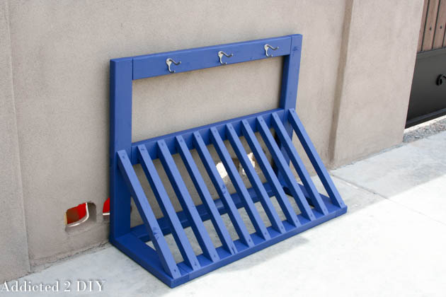 Finished DIY wooden bike rack painted bright blue with coat hooks for helmet storage, leaning against a stucco garage wall