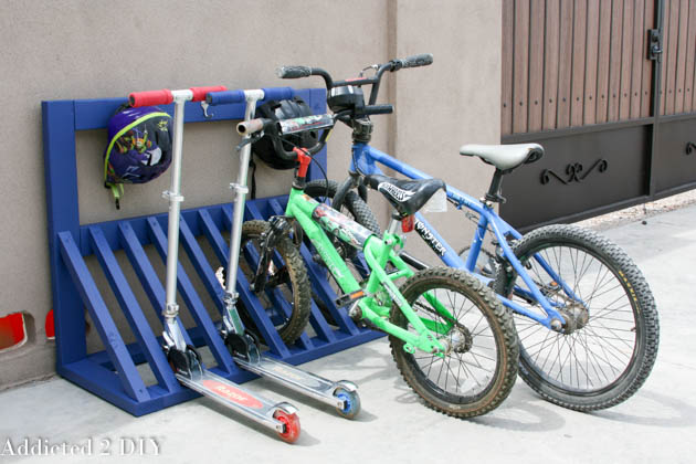 Blue DIY wooden bike rack holding two kids' bicycles, two scooters, and a helmet hung on a coat hook outside a garage