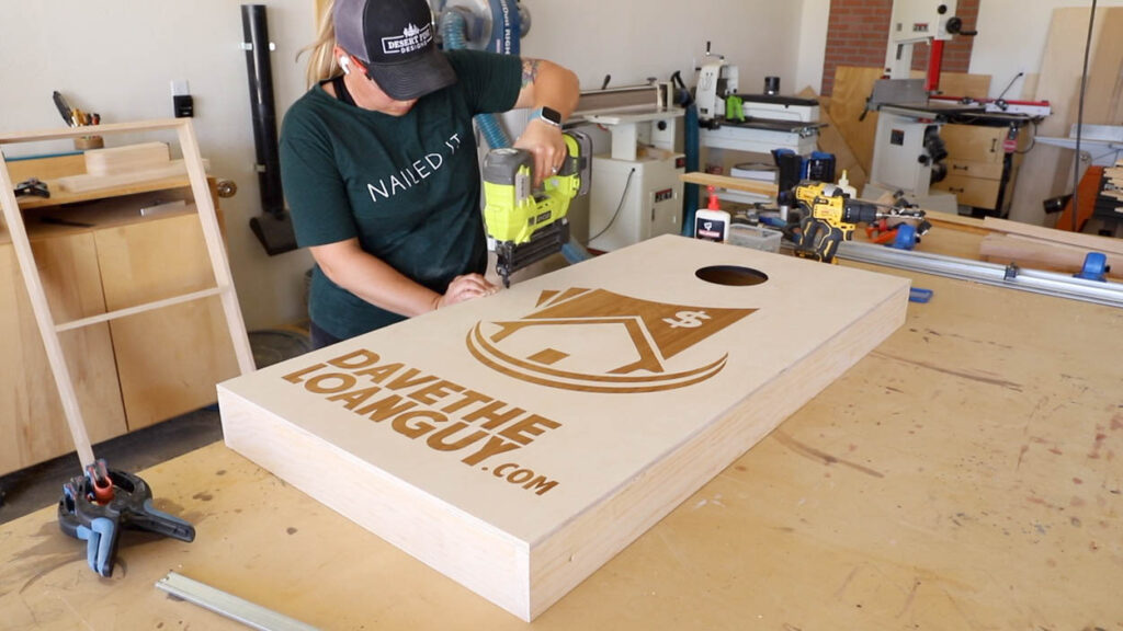 Nailing the laser-engraved plywood cornhole board top to the pine frame with a brad nailer