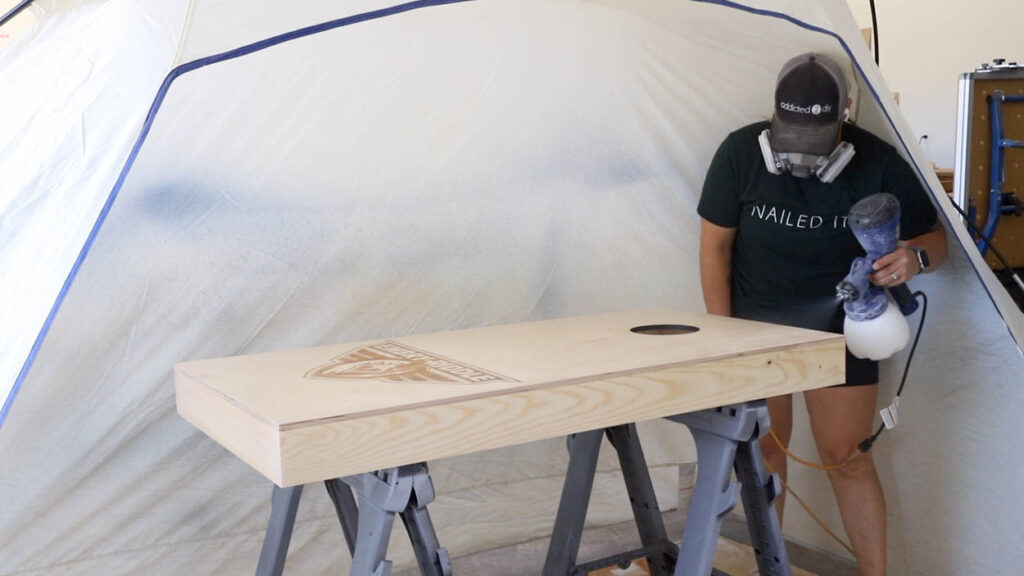 Spraying multiple coats of water-based polyurethane onto a laser-engraved cornhole board using a paint sprayer inside a spray tent