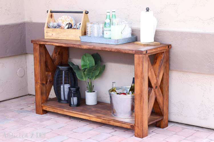 Angled view of finished DIY rustic outdoor console table with X-brace sides, lower shelf, and oil rubbed bronze lag bolts styled with a drink station setup on a brick patio