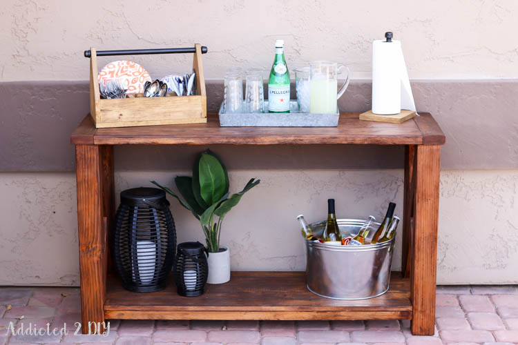 Front view of finished DIY rustic outdoor console table styled with a galvanized drink tray, DIY utensil caddy, lanterns, potted plant, and ice bucket on a covered patio