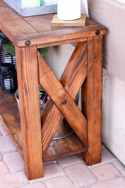 Close-up of X-brace end panel on DIY rustic outdoor console table showing oil rubbed bronze decorative lag bolts on the breadboard end and brace center