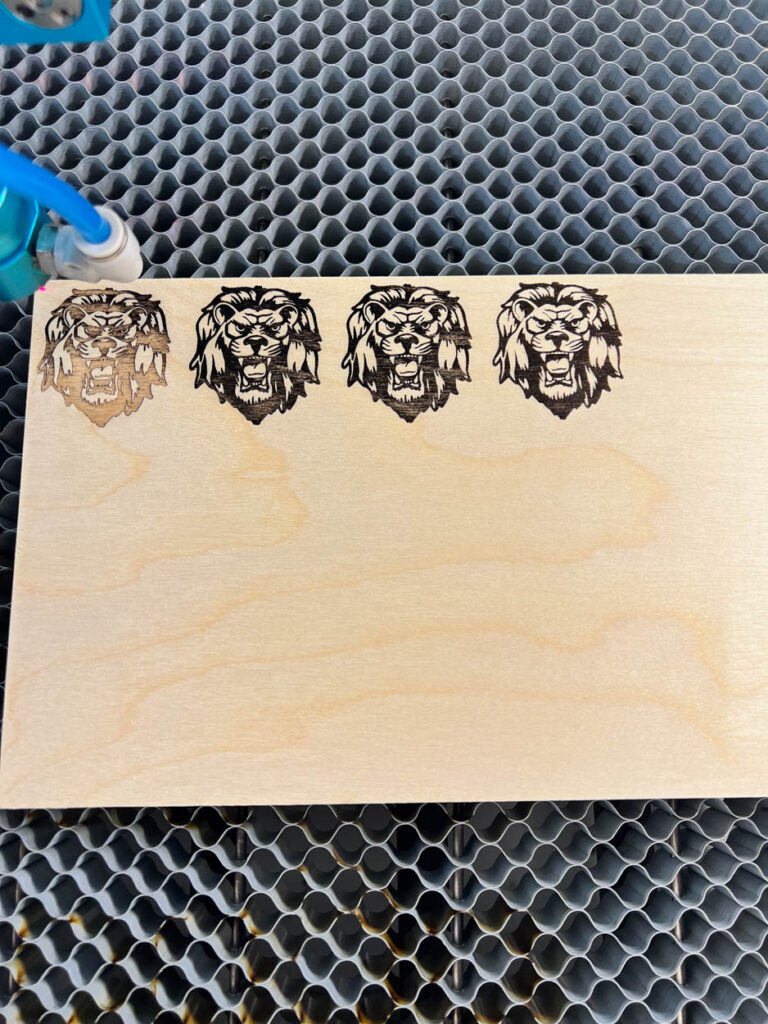 Four lion mascot logos laser engraved in a row on a scrap piece of Baltic birch plywood inside a Thunder Laser, showing the progression of different power and speed settings during the testing process