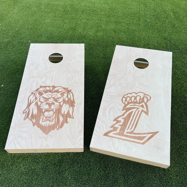 Two DIY cornhole boards with laser-engraved designs on grass — one featuring a detailed lion mascot head, the other a letter logo with crown, both engraved on natural Baltic birch plywood