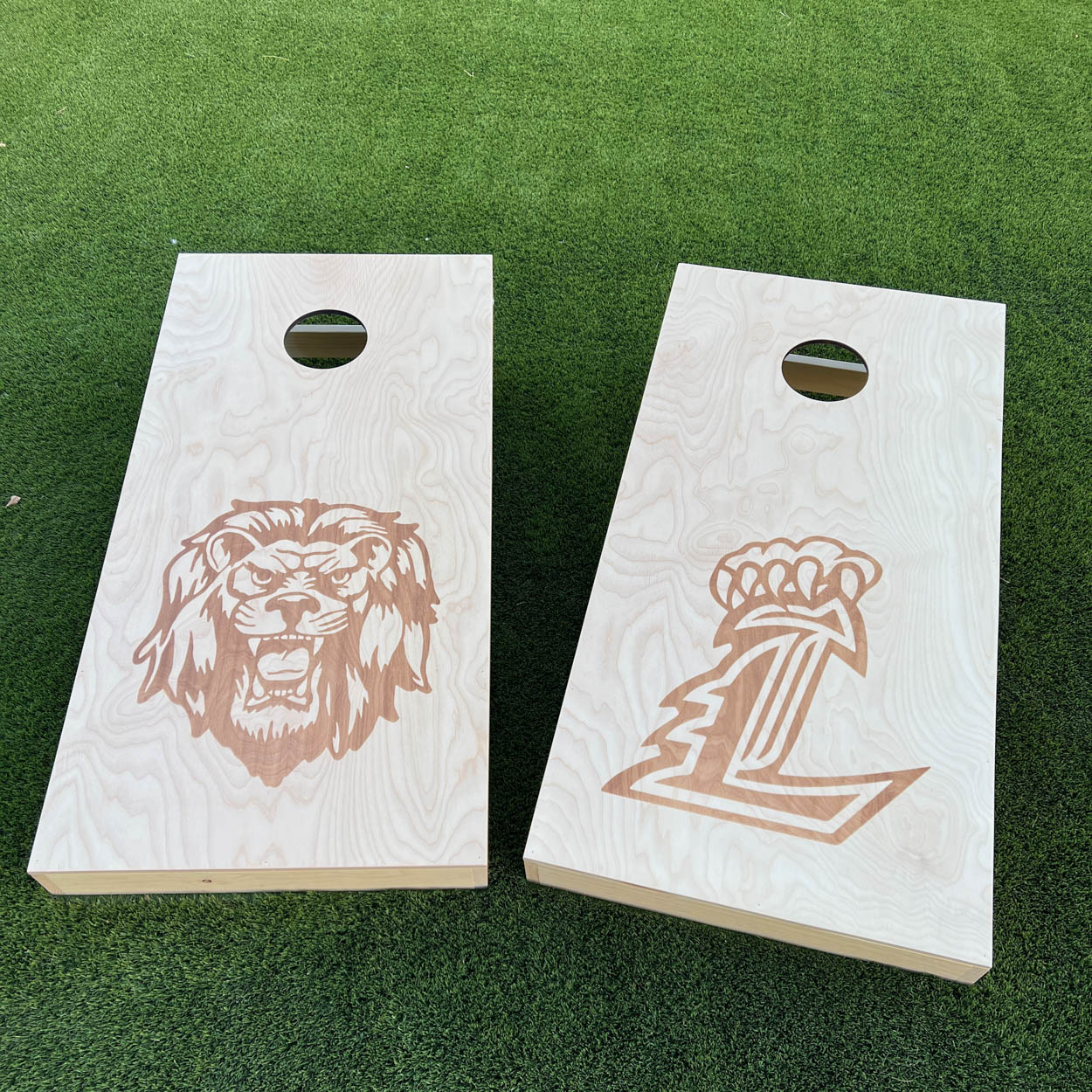Two DIY cornhole boards with laser-engraved designs on grass — one featuring a detailed lion mascot head, the other a letter logo with crown, both engraved on natural Baltic birch plywood