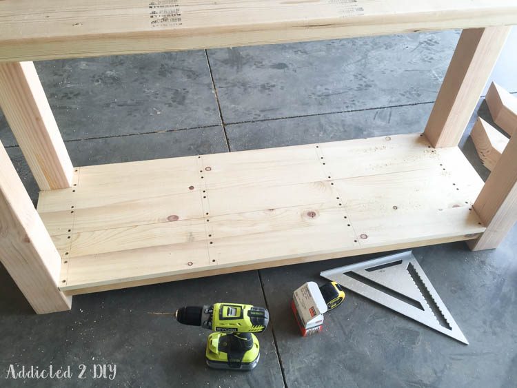 Bottom shelf boards attached to the console table frame with pocket screws, shown with a Ryobi drill and speed square on the workshop floor