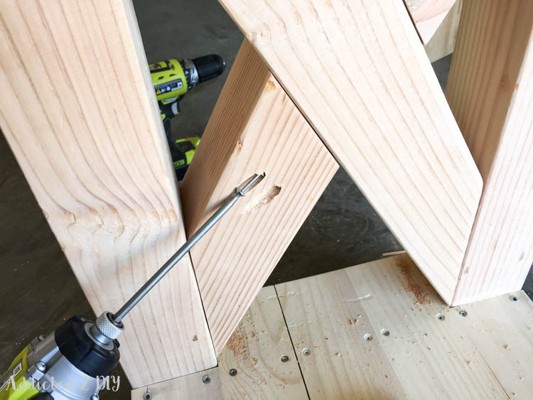 Long drill bit drilling pocket holes into the 30-degree angled center joint of the X-brace on the DIY rustic outdoor console table