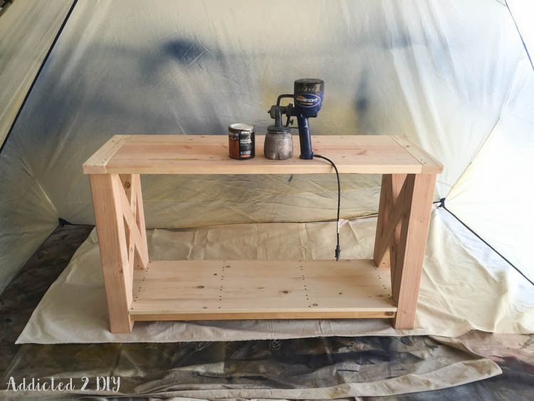 Fully assembled unfinished DIY console table set up in a paint spray shelter with an HVLP sprayer and can of Behr weatherproof stain in natural cedar