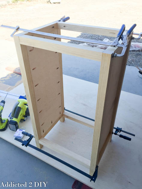Partially assembled DIY wine cabinet frame with one plywood side panel attached, clamped on a workbench outdoors