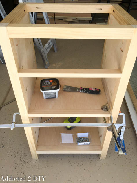 DIY wine cabinet frame with both shelves installed, a container of wood filler and putty knife sitting on the middle shelf