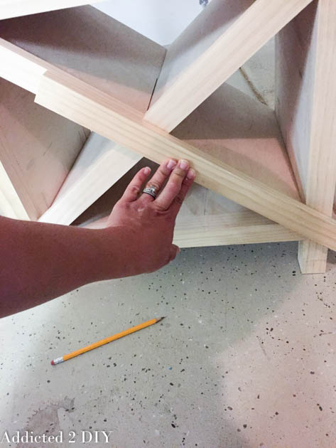 Hand holding a 1x2 trim piece flush against the X wine rack to mark the miter angle with a pencil
