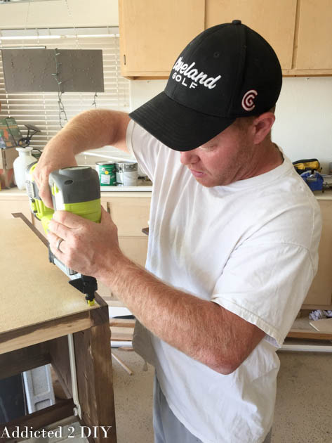 Man using a Ryobi brad nailer to attach the back panel to a stained DIY wine cabinet in a garage workshop