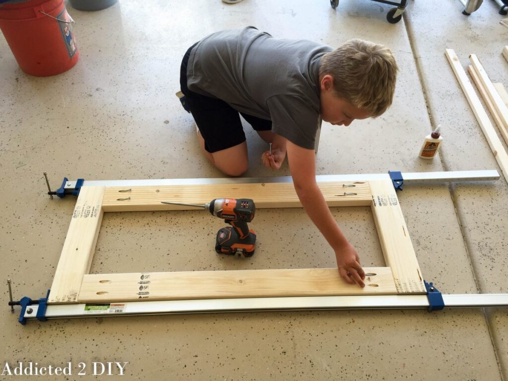 Child helping assemble the rectangular base frame of a DIY bike rack using a drill and clamps on a garage floor