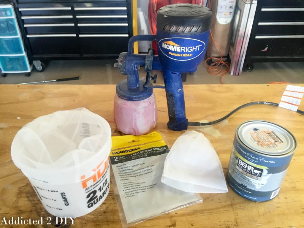 HomeRight Finish Max HVLP paint sprayer, paint strainer bags, mixing bucket, and a can of Behr Premium Plus satin enamel laid out on a workshop bench