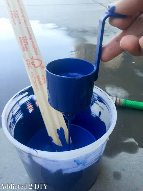 Blue latex paint being measured and diluted with water in a mixing bucket using a paint stick and measuring cup